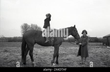 1960er Jahre, historisch, draußen in einer Vorstadtreitschule, ein kleiner Junge, der rückwärts auf einem Pferd sitzt, Hände auf dem Kopf, die Mutter hält die Zügel des Pferdes. So auf einem Pferd zu sitzen, hilft beim Ausbalancieren. Stockfoto