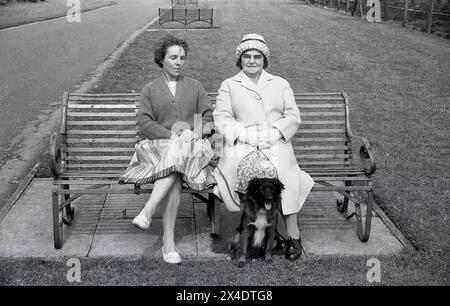 1960er Jahre, historisch, eine Tochter und mit einer älteren Mutter, formell mit Mantel und Hut gekleidet, zusammen auf einer Metallparkbank mit ihrem Hund sitzen, nachdem sie einen Spaziergang gemacht haben i Stockfoto