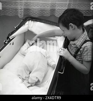 1960er Jahre, historisch, in einem Zimmer, ein kleiner Junge, der die kleine Hand seines kleinen Bruders berührt, der auf dem Rücken liegt in einem Kinderwagen der Epoche. Stockfoto
