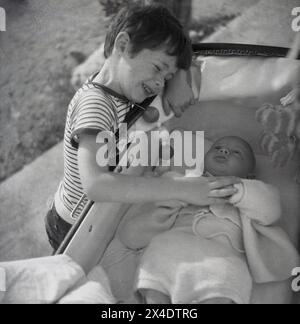 1960er Jahre, historisch, draußen auf einer Terrasse, streichelte ein kleiner Junge die kleinen Hände seines kleinen Bruders, der sich in einem Kinderwagen dieser Zeit auf dem Rücken legt. Stockfoto