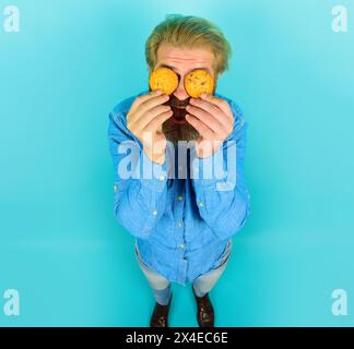 Leckere Leckereien. Ein lächelnder bärtiger Mann hat die Augen mit Haferflockenkeksen bedeckt. Hübscher Mann in Denim-Hemd mit leckeren Keksen. Frühstück oder Mittagessen Stockfoto