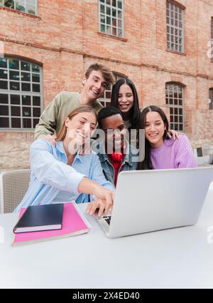 Vertikal. Eine Gruppe multirassischer Teenager-Schüler lächelt mit einem Laptop, um Informationen für die Hausarbeiten der Highschool zu suchen. Klassenkameraden, die für A lernen Stockfoto