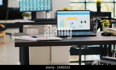 Rollstuhl im Büro, der behinderten Mitarbeitern Zugang bietet, und Laptop auf dem Schreibtisch, der Geschäftsanalysen mit Computerbildschirm im Hintergrund zeigt, auf dem Börsennotierungen angezeigt werden Stockfoto