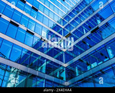 Flacher Blick auf das moderne Bürogebäude aus Glas mit blau getönten Fenstern und geometrischen Architekturmustern, die abstrakte Reflexionen erzeugen. Stockfoto