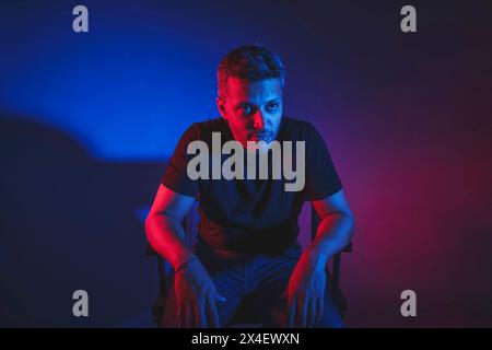 Ein Mann in einem schwarzen Hemd sitzt auf einem Stuhl vor einem blauen Hintergrund Stockfoto