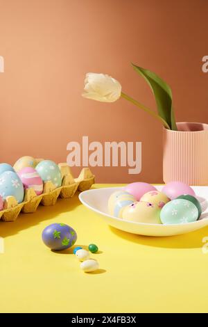 Osterfeier-Konzept mit Blister und weißer Schale mit bunten Eiern, dekoriert mit Blume und Vase auf braunem Hintergrund Stockfoto