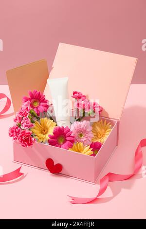 Eine weiße Kosmetikröhre, die auf rosafarbener Geschenkbox voller frischer Blumen mit brauner Papierkarte und rosafarbenem Band auf süßrosa Hintergrund dargestellt wird. Mocku Stockfoto