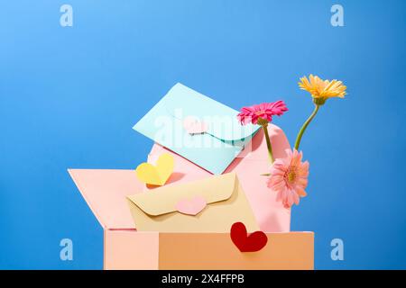 Vorderansicht einer Geschenkbox mit niedlichen handgefertigten Umschlägen und frischen Gerberas auf blauem Hintergrund. Muttertag oder Frauentag Stockfoto