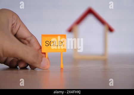 Ein Nahaufnahme-Foto einer Hand mit einem leuchtend orangefarbenen „VERKAUFT“-Schild vor einem hölzernen Miniaturhaus. Immobilien- und Immobilienkonzept. Stockfoto