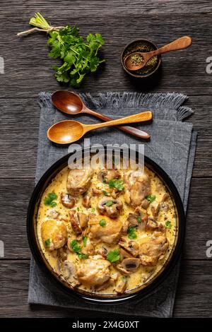 Hähnchenschenkel in einer cremigen Pilz-Knoblauchsauce mit Kräutern und Parmesan in Backform auf dunklem Holztisch mit Holzlöffeln und Serviette, ver Stockfoto