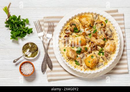 Hähnchenschenkel in einer cremigen Pilz-Knoblauchsauce mit Kräutern und Parmesan in weißer Auflaufform auf weißem Holztisch mit Gabeln, horizontale Ansicht Stockfoto