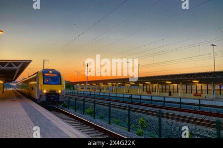 Antwerpen Noorderkempen, Brecht, Antwerpen, 15. Mai 2019, dieses atemberaubende Bild zeigt einen Pendlerzug am Bahnhof Noorderkempen bei einem atemberaubenden Sonnenuntergang. Der Himmel in Orange- und Blautönen strahlt ein warmes Leuchten über die elegante moderne Architektur des Bahnhofs und die übersichtlichen Gleise aus. Die Szene ist frei von Menschen, was die Bereitschaft des Zuges zum Einsteigen unterstreicht, wenn der Tag in die Nacht übergeht, und eine ruhige und dennoch dynamische Momentaufnahme des täglichen Transitverkehrs an einem bestimmten Ort bietet. Sonnenuntergang am Bahnhof Noorderkempen: Pendlerzug unter einem lebhaften Abendhimmel. Hochwertige Fotos Stockfoto