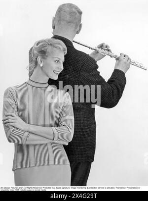 In den 1950er Jahren Eine junge Frau, die von vorne gesehen wird und zuhört, während ein Mann, der von hinten abgebildet wird, eine Querflöte spielt. 1956 Stockfoto