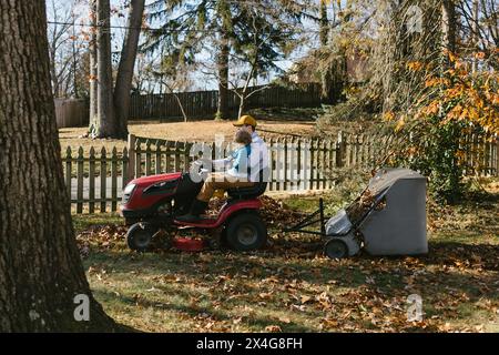 Vater fährt den Rasenmäher mit Kleinkindern, die im Herbst Blätter sammeln Stockfoto