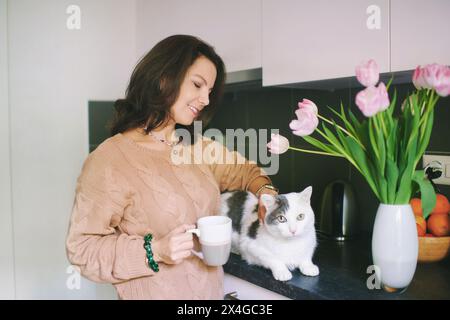 Innenporträt einer hübschen jungen Frau, die mit einer Katze kuschelt und eine Tasse Tee oder Kaffee hält Stockfoto