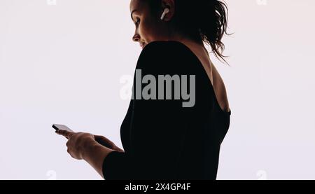 Junge Frau, die in einem Studio steht und ihr Smartphone benutzt, um Musik von einer mobilen App zu streamen. Frauen hören Musik mit schnurlosen Ohrhörern. Stockfoto