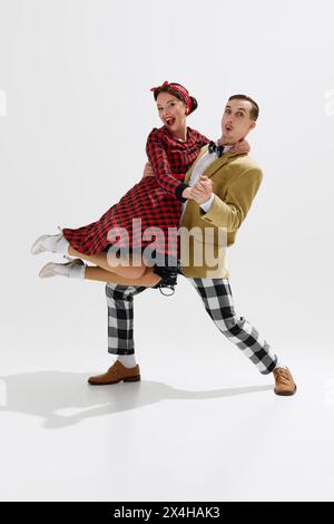 Unterhaltsame und energiegeladene Retro-Tanzszene mit Mann und Frau in farbenfroher Mode der 1950er Jahre, die einen fröhlichen Moment im weißen Studio teilen Stockfoto