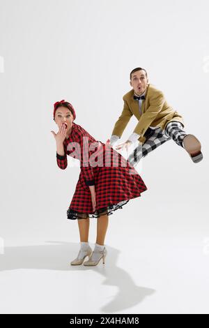 Unterhaltsame und energiegeladene Retro-Tanzszene mit Mann und Frau in farbenfroher Mode der 1950er Jahre, die einen fröhlichen Moment im weißen Studio teilen Stockfoto