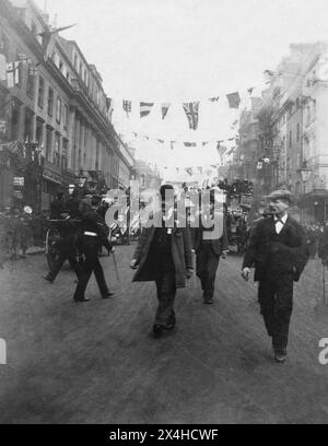 London. 1900: Eine geschäftige Straßenszene in London. Die Straße ist mit Fahnen und Fahnen für die Prozession geschmückt, die am 2. Oktober 1900 zum Gedenken an die Rückkehr der City of London Imperial Volunteers stattfand, die während des Zweiten Burenkrieges in Südafrika gedient hatten. Omnibusse mit Pferden, ein Soldat und zivile Fußgänger sind unterwegs. Ein Mann im Vordergrund trägt einen Bowler-Hut und eine Medaille auf seiner Weste. Stockfoto