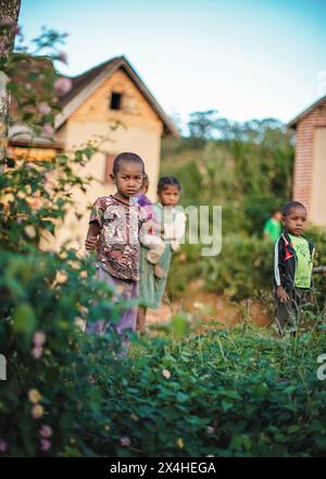 Ankafina-Tsarafidy, Madagaskar - 26. April 2019: Lokaler madagassischer schüchterner Junge, der vor einem einfachen Haus steht, grüne Büsche in der Nähe, weitere Kinder in bac Stockfoto