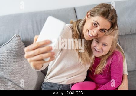 Fröhliche Mutter und Tochter kuscheln und machen ein Selfie auf einer Couch in gemütlicher Atmosphäre Stockfoto