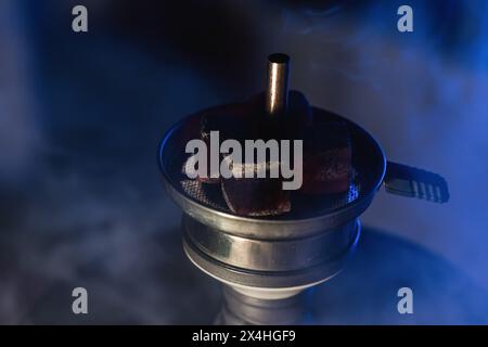 Nahaufnahme glühender Kohlen auf aromatischem Tabak in einer Shisha-Schüssel, die Rauch in einer dunklen, stimmungsvollen Umgebung in einer traditionellen Shisha Lounge im Nahen Osten ausstrahlt. Stockfoto
