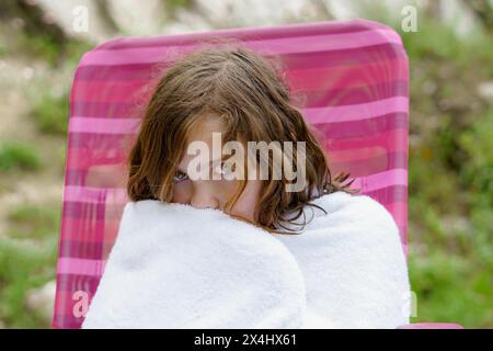 Ein hübsches achtjähriges Mädchen, das auf einem Stuhl sitzt, in ein weißes Handtuch gewickelt, nachdem es aus dem Wasser kam und kalt aussieht Stockfoto