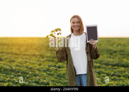 Die lächelnde Agronomin hält ein digitales Tablet mit Mockup-Bildschirm und Sojapflanze im Feld, untersucht, überprüft, schaut in die Kamera. Smart Farming so Stockfoto