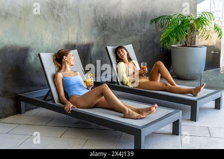 Zwei Freundinnen lachen und trinken Saft im Poolbereich eines schicken Hotels Stockfoto