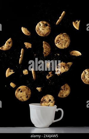 Weißer Becher mit Schokoladenkeksen, die auf schwarzem Hintergrund fliegen Stockfoto