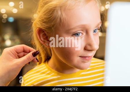 Mädchen lächelt, während sie einen Ohrring mit Hilfe eines Erwachsenen in einem Juweliergeschäft anprobiert Stockfoto