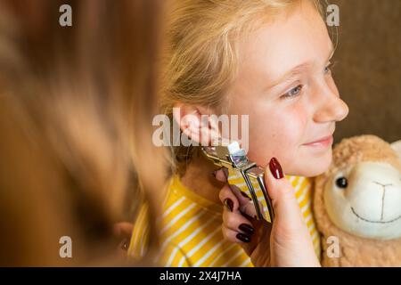 Lächelndes Mädchen, das einen Teddybären beim Ohrpiercing hält, mit Piercingpistole, die von einem professionellen Arzt in einem Schönheitszentrum benutzt wird Stockfoto