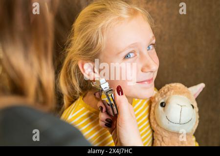 Ein junges Mädchen, das einen Teddybären hält und in die Kamera lächelt, während es in einem Juweliergeschäft von einem professionellen Mediziner ihr Ohr durchbohrt Stockfoto