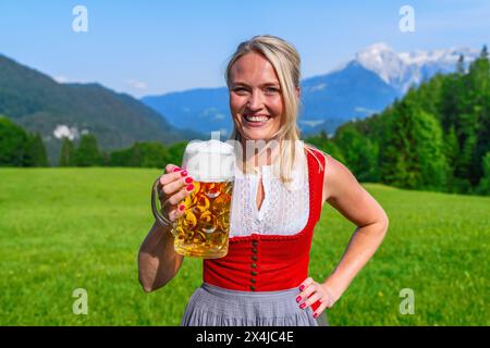 Lächelnde blonde Frau in roter tracht, die auf dem bayerischen oktoberfest mit einem schäumenden Bierkrug mit alpinen Bergen im Hintergrund tobt Stockfoto