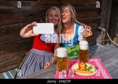 Fröhliche Freundinnen machen Selfie mit Bierkrügen vor einer Almhütte in den bayerischen alpen in traditionellen Dirndls oder tracht. Oktoberfest Bava Stockfoto