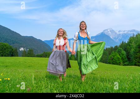 Fröhliche Freundinnen in traditionellen bayerischen Kleidern, Dirndl oder tracht Hände halten und auf einer Sommerwiese auf der deutschen Almwelt herumwirbeln Stockfoto