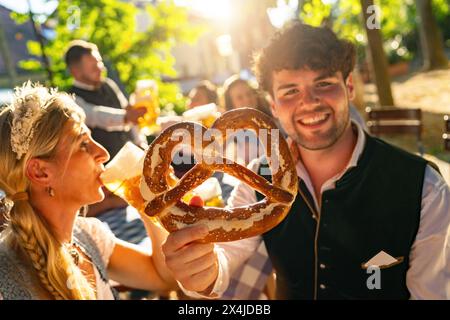 Glückliches Paar mit Bier und großer Brezel auf dem Oktoberfest, traditioneller Kleidung, sunflare Hintergrund Stockfoto