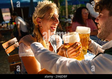 Ein paar lachende und toastende Bierbecher auf dem Oktoberfest, mit goldenem Hinsenlicht und verschwommenem Hintergrund in bayern Stockfoto