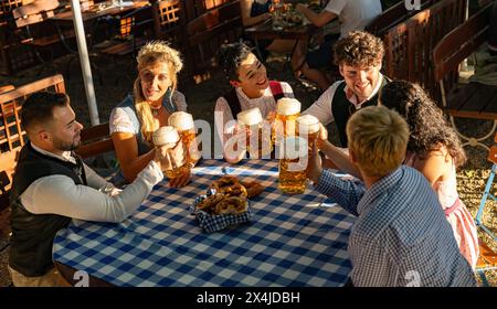 Eine Gruppe von Freunden genießt beim Oktoberfest Bier und Brezeln in traditionellen bayerischen Outfits, Sonnenuntergang Stockfoto