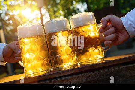 Hände halten bayerische Bierbecher mit Sonnenlicht auf einem Holztisch im Biergarten oder auf dem oktoberfest in Deutschland Stockfoto