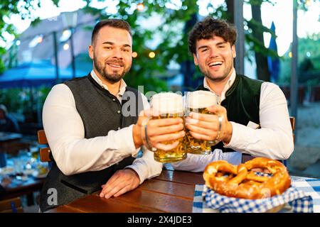 Zwei Männer im traditionellen bayerischen Biergarten oder oktoberfest, die mit Bierbecher an einem Tisch mit Brezel in Bayern anstoßen Stockfoto