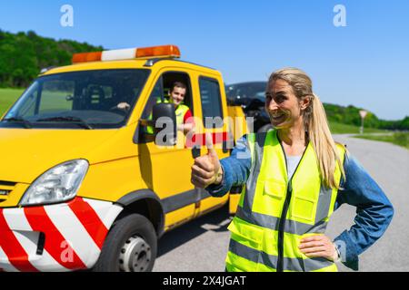 Frau, die die Daumen nach einer Panne mit Abschleppwagen und Fahrer im Hintergrund zeigt. Bild Pannenhilfe-Konzept. Stockfoto