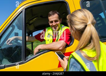 Frau, die ein Smartphone benutzt und mit einem lächelnden Fahrer eines Abschleppwagens spricht, der in einem gelben Lkw sitzt. Bild Pannenhilfe-Konzept. Stockfoto