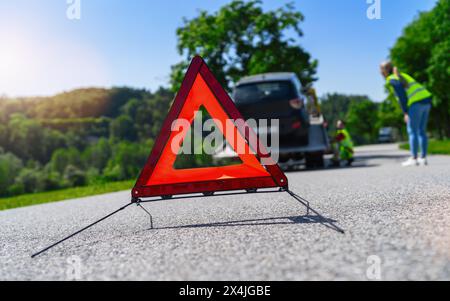 Rotes Warndreieck auf der Straße mit Frau und Abschleppwagen im Hintergrund, die sein signalisierendes Auto überprüfen, das Probleme in einer ländlichen Gegend hat Stockfoto