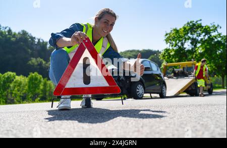 Frau, die ein Warndreieck auf der Straße aufbaut und ist glücklich und zufrieden mit einem schnellen und zuverlässigen Abschleppdienst für Hilfe auf der Straße. Sie zeigt do Stockfoto