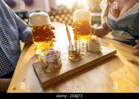 Bayerische Obatzda mit Brezeln und Radieschen und Bierbecher, Mann und junge Frau in tracht im Hintergrund beim Biergarten oder oktoberfest, München, Keim Stockfoto