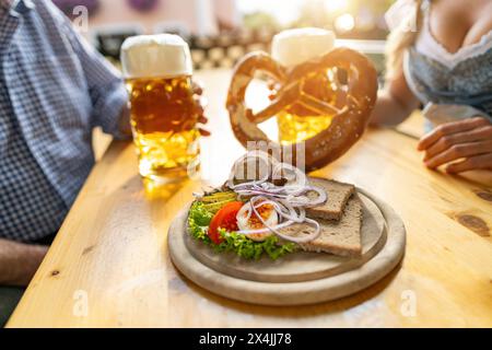 Bayerische Obatzda mit Brezeln und Radieschen und Bierbecher, Mann und junge Frau in tracht im Hintergrund beim Biergarten oder oktoberfest, München, Keim Stockfoto