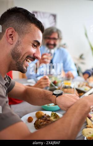 Seitenansicht fröhlicher kaukasischer Mann, der Essen isst, am Tisch sitzend, mit Familie im Hintergrund unfokussiert. Stockfoto