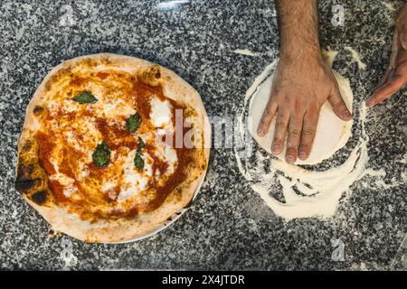 Top-down-Schuss der Hände, die Pizzateig zubereiten, neben vorbereiteter margherita-Pizza, Bäckerei. Hochwertige Fotos Stockfoto