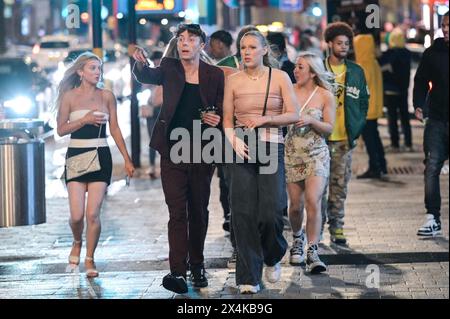 Broad Street, Birmingham, 4. Mai 2024 - am Freitagabend besuchten die Revellers den berüchtigten Nachtclub von Birmingham, um die Feiertage Anfang Mai zu beginnen. Der Regen hat die Partygäste nicht davon abgehalten, sich mit Mänteln oder Handtaschen zu amüsieren, um ihre Haare vor den Elementen zu schützen. Zwei Jungs saßen an einer Bushaltestelle mit einem Pappausschnitt von Louis Tomlinson, den sie aus einem Nachtclub in der Nähe hatten. Der Nachtclub Rosies war besonders voll mit Warteschlangen, die sich um Barrieren schlängelten. Zwei spärlich bekleidete Feuerschauer unterhielten Passanten im Walkabout und die Pubbesucher genossen Musik in Heidi's Bier Bar, während sie tanzten Stockfoto